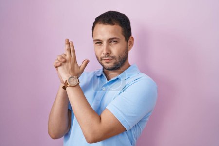 Téléchargez les photos : Homme hispanique debout sur fond rose tenant un pistolet symbolique avec geste de la main, jouant à tuer des armes à feu, visage en colère - en image libre de droit