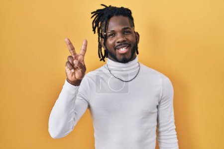 Foto de Hombre africano con rastas con jersey de cuello alto sobre fondo amarillo mostrando y señalando con los dedos número dos mientras sonríe confiado y feliz. - Imagen libre de derechos