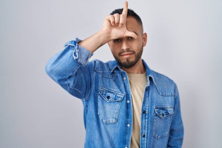 Photo for Young hispanic man standing over isolated background making fun of people with fingers on forehead doing loser gesture mocking and insulting. - Royalty Free Image