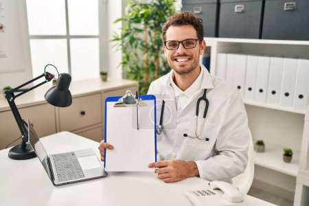 Téléchargez les photos : Jeune homme hispanique portant un stéthoscope médecin tenant presse-papiers regardant positif et heureux debout et souriant avec un sourire confiant montrant des dents - en image libre de droit