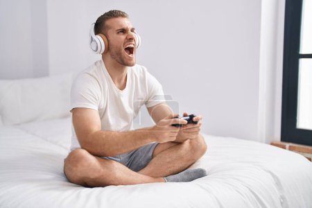 Photo for Young caucasian man sitting on the bed at home playing video games angry and mad screaming frustrated and furious, shouting with anger looking up. - Royalty Free Image