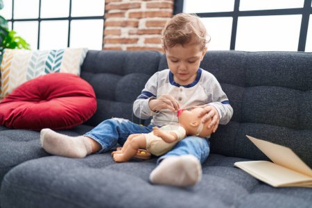 Photo for Adorable caucasian boy playing with baby doll sitting on sofa at home - Royalty Free Image