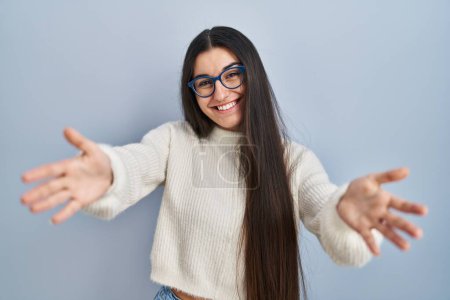 Photo for Young hispanic woman wearing casual sweater over blue background looking at the camera smiling with open arms for hug. cheerful expression embracing happiness. - Royalty Free Image
