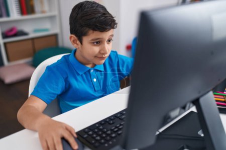 Téléchargez les photos : Adorable élève hispanique garçon en utilisant l'ordinateur assis sur la table en classe - en image libre de droit