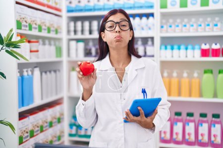 Photo for Young hispanic woman working at pharmacy drugstore holding red heart puffing cheeks with funny face. mouth inflated with air, catching air. - Royalty Free Image