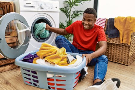 Foto de Young african man putting dirty clothes into washing machine at laundry room - Imagen libre de derechos
