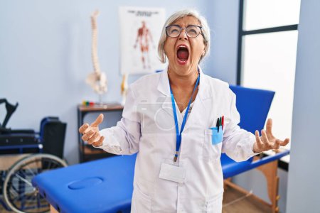 Foto de Middle age woman with grey hair working at pain recovery clinic angry and mad screaming frustrated and furious, shouting with anger. rage and aggressive concept. - Imagen libre de derechos