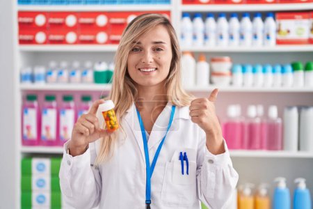 Téléchargez les photos : Young blonde woman working at pharmacy drugstore holding pills smiling happy and positive, thumb up doing excellent and approval sign - en image libre de droit