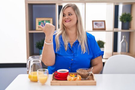 Téléchargez les photos : Caucasian plus size woman eating breakfast at home smiling with happy face looking and pointing to the side with thumb up. - en image libre de droit