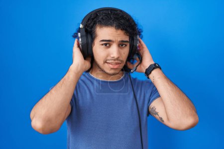Foto de Hispanic man with curly hair listening to music using headphones covering ears with fingers with annoyed expression for the noise of loud music. deaf concept. - Imagen libre de derechos