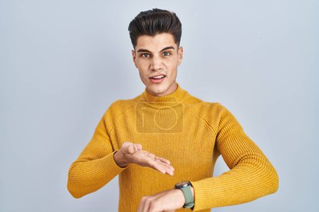 Téléchargez les photos : Jeune homme hispanique debout sur fond bleu à la hâte pointant pour regarder le temps, l'impatience, bouleversé et en colère pour retard de délai - en image libre de droit