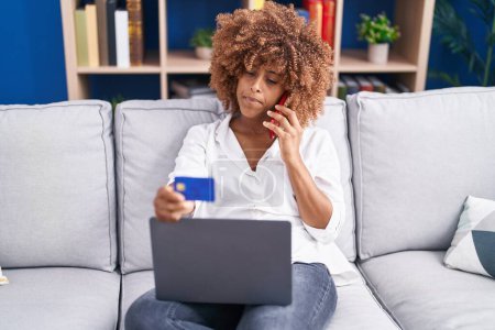 Téléchargez les photos : Femme afro-américaine parlant sur smartphone tenant la carte de crédit assis sur le canapé à la maison - en image libre de droit