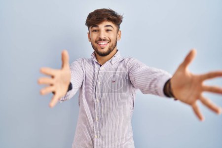 Téléchargez les photos : Arab man with beard standing over blue background looking at the camera smiling with open arms for hug. cheerful expression embracing happiness. - en image libre de droit