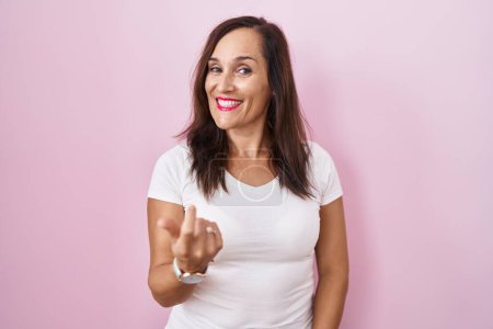 Foto de Middle age brunette woman standing over pink background beckoning come here gesture with hand inviting welcoming happy and smiling - Imagen libre de derechos