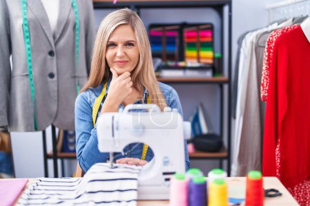 Foto de Blonde woman dressmaker designer using sew machine looking confident at the camera smiling with crossed arms and hand raised on chin. thinking positive. - Imagen libre de derechos