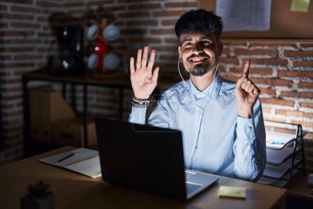 Foto de Joven hombre hispano con barba trabajando en la oficina por la noche mostrando y señalando con los dedos el número seis mientras sonríe confiado y feliz. - Imagen libre de derechos
