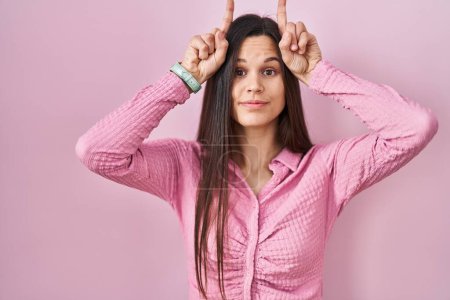 Téléchargez les photos : Jeune femme hispanique debout sur fond rose faisant un geste drôle avec le doigt sur la tête comme des cornes de taureau - en image libre de droit