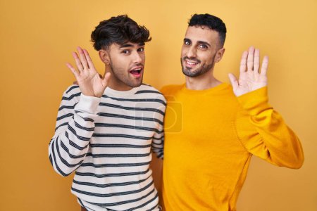 Téléchargez les photos : Jeune couple gay hispanique debout sur fond jaune renonçant à dire bonjour heureux et souriant, geste de bienvenue amical - en image libre de droit