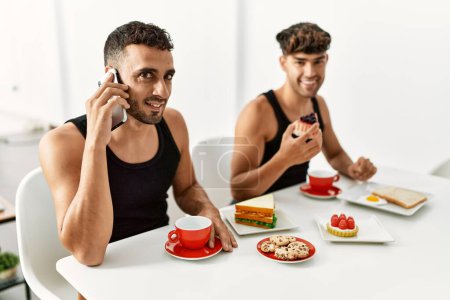 Foto de Two hispanic men couple talking on the smartphone having breakfast at home - Imagen libre de derechos