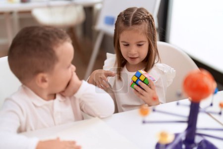 Foto de Adorable chica y niño jugando con el cubo de rompecabezas de color sentado en la mesa en el jardín de infantes - Imagen libre de derechos