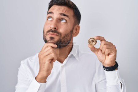 Foto de Handsome hispanic man holding polkadot cryptocurrency coin serious face thinking about question with hand on chin, thoughtful about confusing idea - Imagen libre de derechos