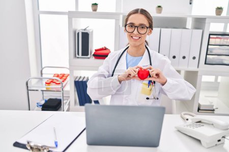 Photo for Young beautiful hispanic woman doctor using laptop working holding heart at clinic - Royalty Free Image