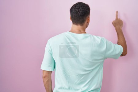 Téléchargez les photos : Bel homme hispanique debout sur fond rose posant vers l'arrière pointant vers l'avant avec la main du doigt - en image libre de droit