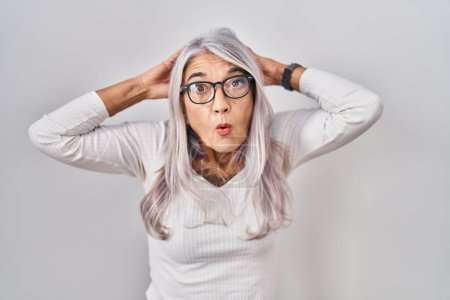 Foto de Middle age woman with grey hair standing over white background crazy and scared with hands on head, afraid and surprised of shock with open mouth - Imagen libre de derechos