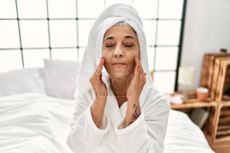 Foto de Mujer de pelo gris de mediana edad masajeando la cara con crema sentada en la cama en el dormitorio - Imagen libre de derechos