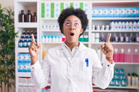 Téléchargez les photos : Belle femme africaine aux cheveux bouclés travaillant à la pharmacie droguerie étonnée et surprise regardant vers le haut et pointant avec les doigts et les bras levés. - en image libre de droit
