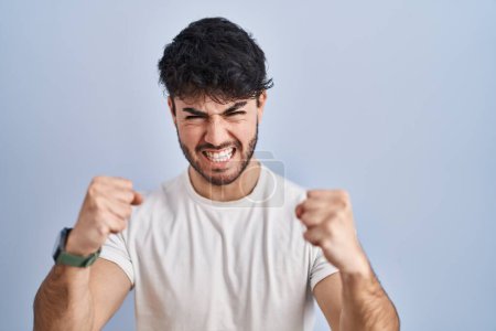 Téléchargez les photos : Homme hispanique avec la barbe debout sur fond blanc en colère et fou levant les poings frustrés et furieux tout en criant avec colère. rage et concept agressif. - en image libre de droit