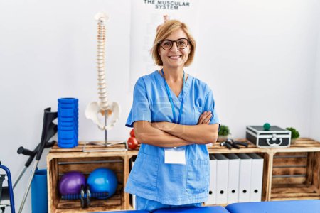 Photo for Middle age blonde woman wearing physio therapy uniform standing with arms crossed gesture at clinic - Royalty Free Image