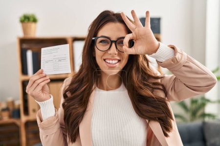 Téléchargez les photos : Jeune femme brune tenant une carte de disque covid souriant heureux faisant signe ok avec la main sur les yeux regardant à travers les doigts - en image libre de droit