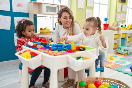 Foto de Profesor con niñas jugando con bloques de construcción sentado en la mesa en el jardín de infantes - Imagen libre de derechos
