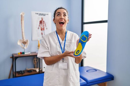 Foto de Mujer hispana joven sosteniendo plantilla de zapato en clínica de fisioterapia enojada y loca gritando frustrada y furiosa, gritando con rabia mirando hacia arriba. - Imagen libre de derechos