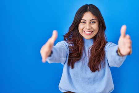Téléchargez les photos : Hispanic young woman standing over blue background looking at the camera smiling with open arms for hug. cheerful expression embracing happiness. - en image libre de droit