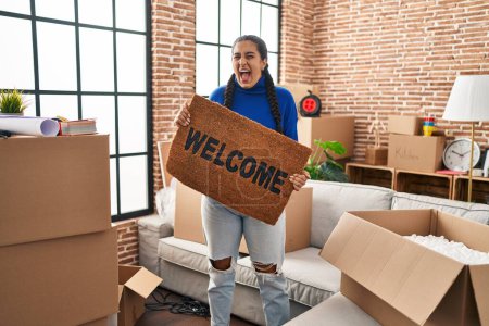 Photo for Young hispanic woman holding welcome doormat at new home angry and mad screaming frustrated and furious, shouting with anger. rage and aggressive concept. - Royalty Free Image