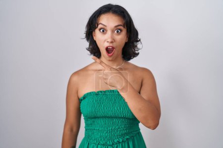 Foto de Young hispanic woman standing over isolated background surprised pointing with finger to the side, open mouth amazed expression. - Imagen libre de derechos