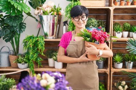 Foto de Joven mujer asiática con pelo corto trabajando en floristería sosteniendo planta mirando positiva y feliz de pie y sonriendo con una sonrisa confiada mostrando los dientes - Imagen libre de derechos
