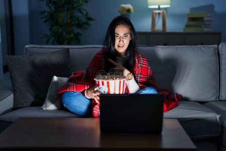 Foto de Hispanic woman eating popcorn watching a movie on the sofa pointing aside worried and nervous with forefinger, concerned and surprised expression - Imagen libre de derechos