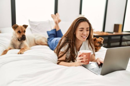Téléchargez les photos : Jeune femme hispanique utilisant un ordinateur portable et buvant du café couché sur le lit avec des chiens dans la chambre - en image libre de droit