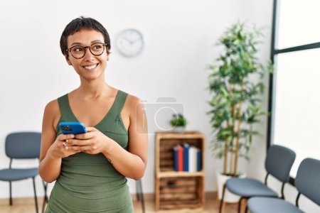 Téléchargez les photos : Jeune femme hispanique souriant confiant en utilisant smartphone debout à la salle d'attente - en image libre de droit