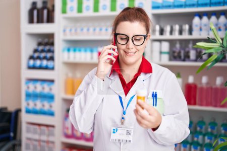 Téléchargez les photos : Jeune belle plus taille femme pharmacien tenant bouteille pilules parler sur smartphone à la pharmacie - en image libre de droit