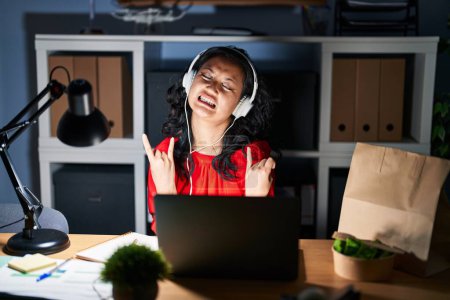 Photo for Young asian woman working at the office with laptop at night shouting with crazy expression doing rock symbol with hands up. music star. heavy concept. - Royalty Free Image