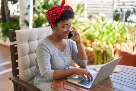Photo for African american woman using laptop and talking on smartphone sitting on table at home terrace - Royalty Free Image