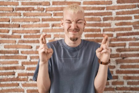 Téléchargez les photos : Jeune homme caucasien debout sur un mur de briques geste doigt croisé souriant avec espoir et les yeux fermés. chance et concept superstitieux. - en image libre de droit