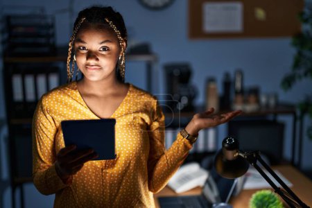 Téléchargez les photos : Femme afro-américaine avec des tresses travaillant au bureau la nuit avec une tablette souriante et joyeuse présentant et pointant la paume de la main en regardant la caméra. - en image libre de droit