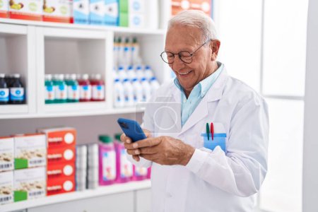 Téléchargez les photos : Homme âgé aux cheveux gris pharmacien utilisant un smartphone à la pharmacie - en image libre de droit