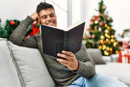 Photo for Young hispanic man reading book sitting on sofa by christmas tree at home - Royalty Free Image