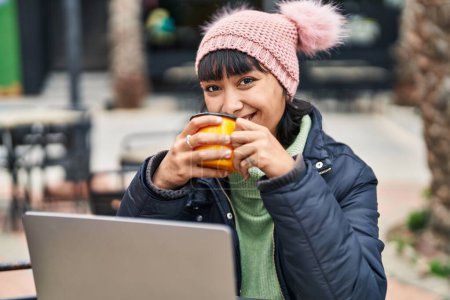 Photo for Young beautiful hispanic woman using laptop drinking coffee sitting on table at coffee shop terrace - Royalty Free Image